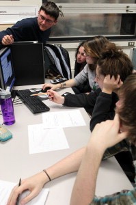 A group of five students surround a lab room desktop computer, clearly collaborating in their learning.