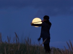 A person stands in twilight in a field, measuring the size of the Moon from their perspective with a measuring tape.