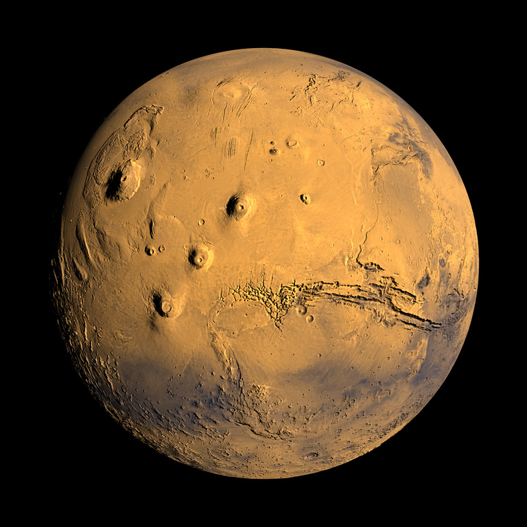 Mars is seen; the planet is orange-brown with patches of black-brown, especially in the south. Mountains, canyons, and craters are visible; mountains at center left and a canyon at center right are notably large.