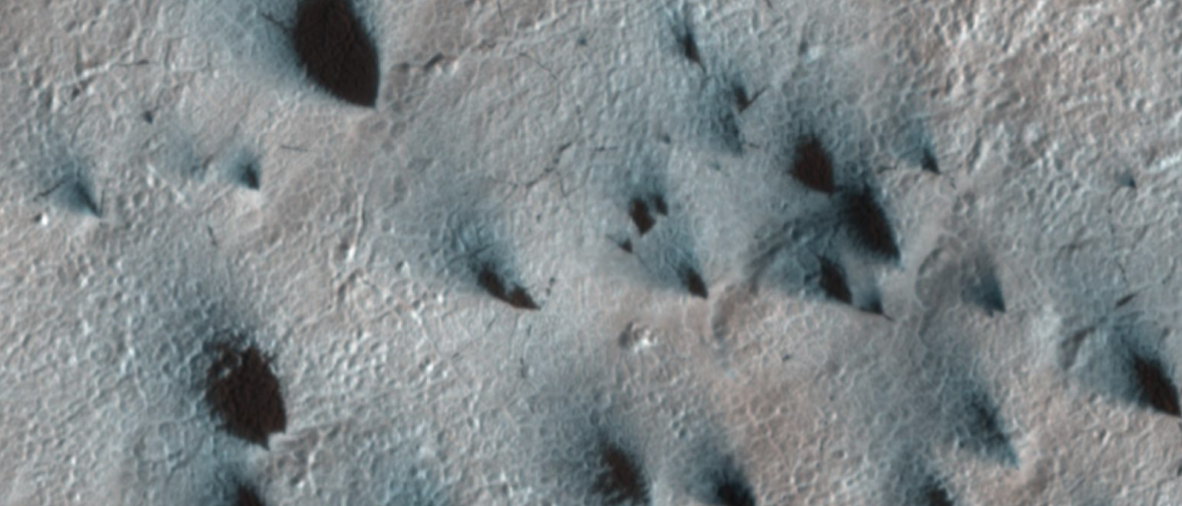 Inky black-blue fan-shapes stretch across a grey surface from bottom right to top left: dust from Martian gas-geyser activity.