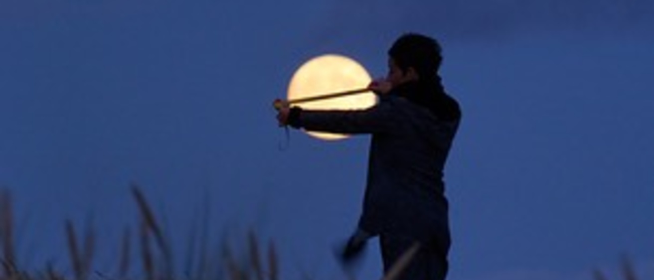 A person stands in twilight in a field, measuring the size of the Moon from their perspective with a measuring tape.